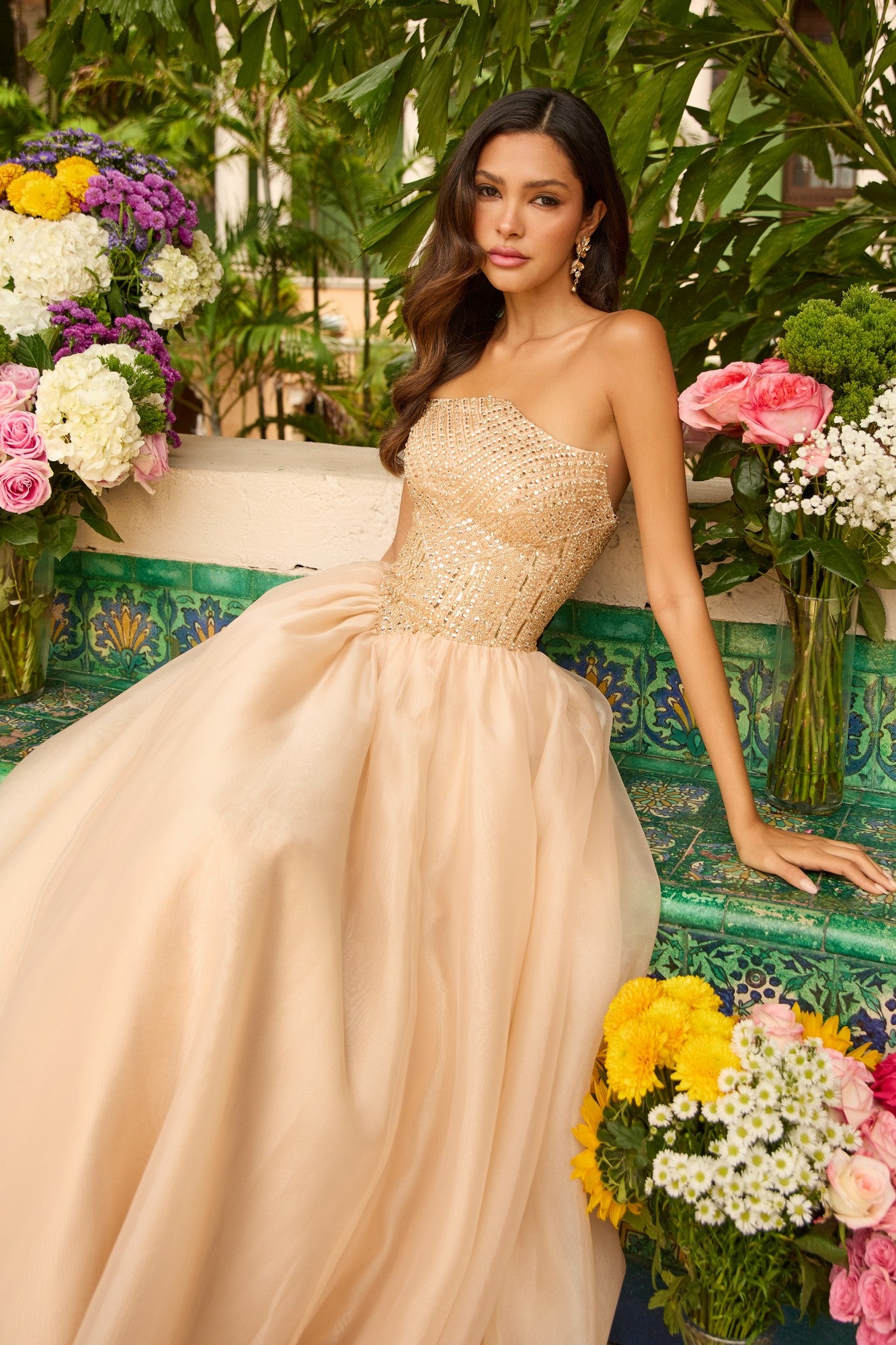 A woman in the Ava Presley 47255 Long Prom Dress, a strapless ball gown with an embellished bodice, sits on a tiled bench amid colorful flowers and lush greenery.